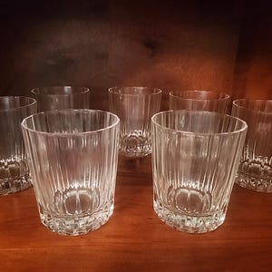 May include: Set of seven clear, ribbed-design drinking glasses. The glasses are cylindrical with a slightly flared top and a patterned base. They are arranged on a dark wooden surface, suggesting a bar or dining setting.