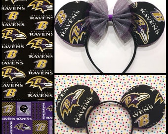 May include: Black and purple Baltimore Ravens themed mouse ears headband. The ears are round with the team logo and text. A purple bow with a silver mesh overlay sits between the ears. The headband is black.