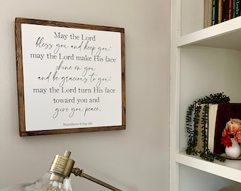 May include: A square wooden wall sign with a dark brown frame and a white background. The sign features black printed text that reads: May the Lord bless you and keep you: may the Lord make His face shine on you and be gracious to you: may the Lord turn His face toward you and give you peace. Numbers 6:24-26.