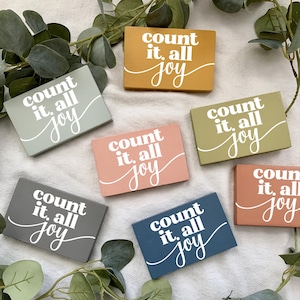 May include: Six wooden signs with the text "count it, all joy" in white lettering on a variety of colored backgrounds. The signs are arranged in a semi-circle on a white surface.