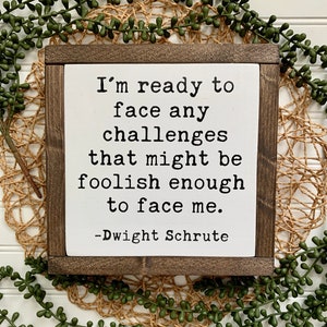 May include: A white wooden sign with a brown frame, featuring a quote from the TV show "The Office" by Dwight Schrute: "I'm ready to face any challenges that might be foolish enough to face me."