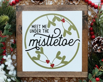May include: A wooden sign with a white background and a green circle with a mistletoe design. The sign reads "Meet me under the mistletoe".