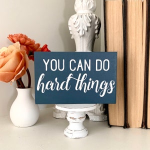 May include: A dark blue wooden sign with white text that reads "You can do hard things". The sign is sitting on a small white pedestal.
