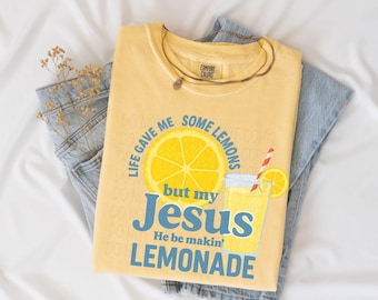 May include: A yellow Comfort Colors t-shirt with the text "Life gave me some lemons but my Jesus He be makin' lemonade" and a graphic of a lemon and lemonade. The shirt is folded on top of blue jeans.