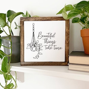 May include: A white wooden sign with a brown frame, featuring a black and white illustration of a hanging plant and the text "Beautiful things take time".