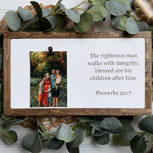 May include: A framed wood sign with a white background and a black clip holding a photo of a family of five. The sign has the text "The righteous man walks with integrity; blessed are his children after him. Proverbs 20:7"