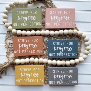 May include: Five wooden signs with the text "STRIVE FOR progress NOT PERFECTION" in white lettering. The signs are painted in different colors: green, pink, orange, blue, and yellow.