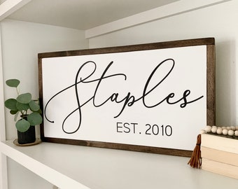 May include: A white wooden sign with a brown frame, featuring the text "Staples EST. 2010" in black cursive lettering.
