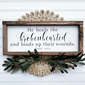 May include: A wooden sign with a white background and black text that reads "He heals the brokenhearted and binds up their wounds. Psalm 147:3". The sign is framed in brown wood and is decorated with a green garland.