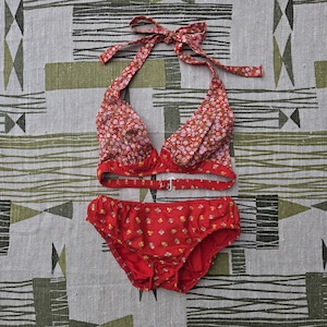 May include: A two-piece swimsuit set featuring a halter top and matching bottoms. The top and bottom are red with a floral pattern in shades of pink, yellow, and brown. The halter top has a tie closure at the neck and a clasp closure at the back.
