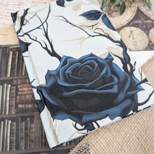 Peut inclure: Une couverture de livre avec une grande rose bleu foncé et des tiges noires sur fond crème. Le motif floral gothique comprend des branches et des feuilles. La couverture rectangulaire semble être en tissu.
