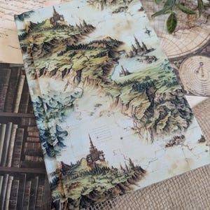 Peut inclure: Un journal à couverture rigide avec un motif de carte vintage détaillé. La carte représente un paysage fantastique avec des montagnes, des forêts et des châteaux dans des tons de vert, de marron et de beige. Le journal est ouvert, révélant les détails complexes de la carte.