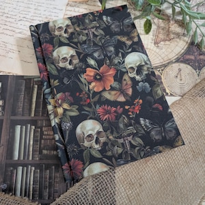 Peut inclure: Un journal sombre avec un motif floral et de crânes. La couverture présente des crânes blancs, des fleurs orange et rouges, et des papillons. Le journal est ouvert, révélant l'intérieur à motifs.