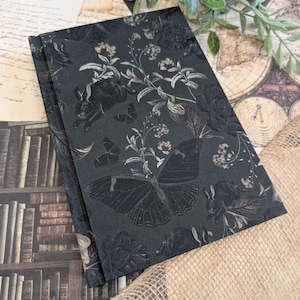 Peut inclure: Deux cahiers gris foncé avec un motif floral et faunique. Le motif comprend des papillons, des lapins et des fleurs dans un motif ton sur ton. Les cahiers sont empilés, celui du dessus étant légèrement incliné, révélant les bords.