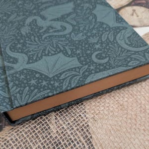 Peut inclure: Un livre à couverture rigide de couleur sarcelle avec un motif de dragon et de fleurs. Les tranches du livre sont de couleur marron clair. Le livre repose sur une surface en toile de jute.