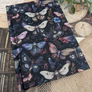 Peut inclure: Un tissu plié de couleur sombre, orné d'un motif répété de papillons de nuit aux couleurs variées, notamment blanc, bleu et rose, ainsi que de motifs floraux. Le design évoque un style d'illustration botanique vintage. Le tissu est plié, mettant en valeur le motif.