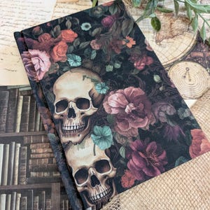 Peut inclure: Un journal à couverture rigide fermé avec un motif floral sombre et des crânes. La couverture présente deux crânes entourés de fleurs bordeaux, roses et turquoise sur fond noir. Le journal est neuf.