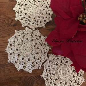 May include: Three white crocheted doilies with a delicate floral pattern. The doilies are arranged on a wooden surface with a red poinsettia flower in the background. The text "Elorne Patterns" is visible in the image.