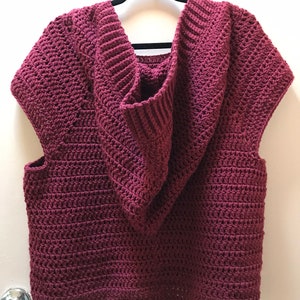 K&ouml;nnte beinhalten: Ein &auml;rmelloser, weinroter H&auml;kel-Hoodie h&auml;ngt an einem schwarzen Kleiderb&uuml;gel. Der Hoodie hat einen tiefen V-Ausschnitt und eine gerippte, &uuml;bergro&szlig;e Kapuze. Der H&auml;kelstich erzeugt ein strukturiertes Aussehen. Das Kleidungsst&uuml;ck wird vor einer wei&szlig;en Wand pr&auml;sentiert.