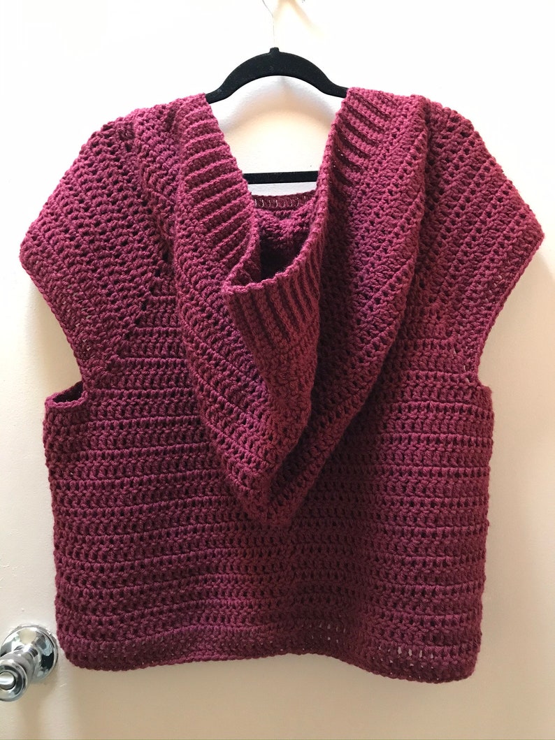 K&ouml;nnte beinhalten: Ein &auml;rmelloser, weinroter H&auml;kel-Hoodie h&auml;ngt an einem schwarzen Kleiderb&uuml;gel. Der Hoodie hat einen tiefen V-Ausschnitt und eine gerippte, &uuml;bergro&szlig;e Kapuze. Der H&auml;kelstich erzeugt ein strukturiertes Aussehen. Das Kleidungsst&uuml;ck wird vor einer wei&szlig;en Wand pr&auml;sentiert.