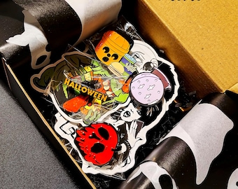 May include: A black box with the word "Mini" printed on the top. The box is filled with Halloween-themed enamel pins, including a ghost, a pumpkin, a skull, and a witch. The pins are packaged in clear plastic bags.