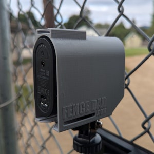 May include: A gray 3D-printed device holder attached to a chain-link fence. The holder encases a black electronic device with visible ports and the text "Fence Dad .com". The device is mounted on a tripod.