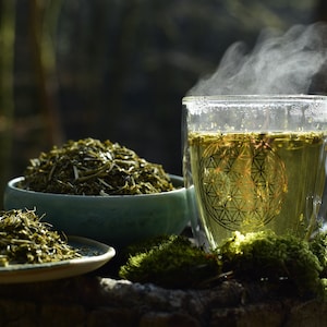 May include: A steaming cup of green tea in a clear glass mug with a geometric design. Loose leaf green tea is shown in a bowl and a small plate.