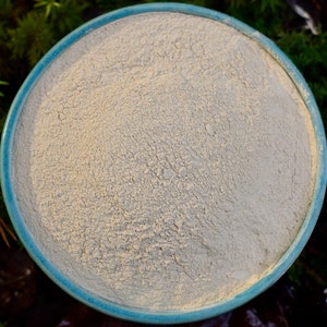 May include: A close-up view of a light beige powder in a blue ceramic bowl. The powder is smooth and fine, with a slightly grainy texture.