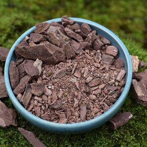 Può includere: Una ciotola in ceramica azzurra riempita con un misto di piccoli frammenti di pietra frastagliati color marrone rossastro e ghiaia fine. Diversi pezzi più grandi, piatti e angolari della stessa roccia marrone rossastra sono sparsi alla base.