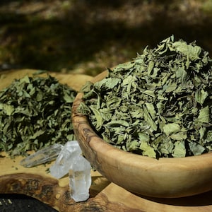 May include: A wooden bowl filled with dried green leaves, accompanied by a separate pile of the same dried leaves and two clear, pointed quartz crystals resting on a natural wood slab with a visible grain and irregular, organic edges.
