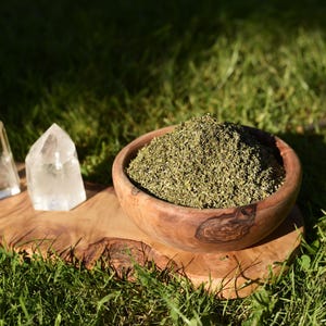 Peut inclure: Un bol en bois rempli d'herbes vertes séchées est posé sur une planche de bois, à côté de deux pointes de cristal de quartz transparent. Les objets sont disposés sur de l'herbe verte, avec des ombres.