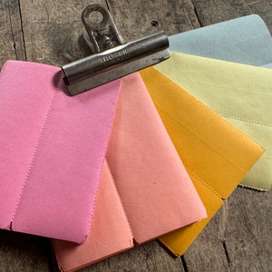 May include: A collection of colorful paper envelopes in shades of pink, peach, yellow, and blue, held together by a silver metal clip. The envelopes are arranged in a fan shape, showcasing their various hues and textures.