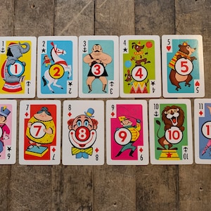 May include: A set of 11 playing cards featuring circus-themed illustrations. Each card has a different number from 1 to 11, with a corresponding circus performer or animal. The cards are colorful and have a vintage style.