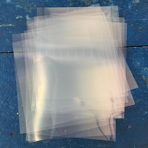 May include: A stack of clear, transparent plastic sheets, likely for protecting documents or photos. The sheets are rectangular and have a slightly frosted appearance. They are stacked on a weathered, blue wooden surface.