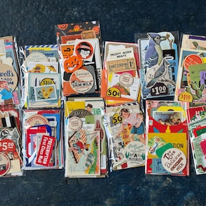 May include: A collection of twelve clear plastic pouches filled with assorted vintage-style paper ephemera, including colorful die-cut labels, price tags, illustrated postcards, dairy coupons, and small printed cards featuring various retro graphics.