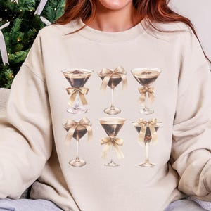 Könnte beinhalten: Beiges Sweatshirt mit einem Aufdruck von sechs Martinigläsern. Jedes Glas enthält eine dunkle Flüssigkeit und ist mit einer goldenen Schleife verziert. Die Gläser sind in zwei Reihen zu je drei angeordnet.