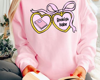 May include: A pink sweatshirt featuring a graphic of a heart-shaped locket with a book inside and the text "Bookish babe".