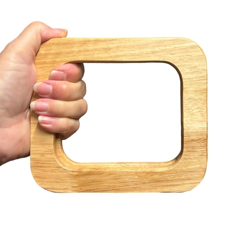 Puede incluir: Un asa de bolso de madera de color claro con forma rectangular y esquinas redondeadas. El asa est&aacute; sujeta por una mano, mostrando la veta natural de la madera y el acabado liso. Dise&ntilde;ado para bolsos y carteras.