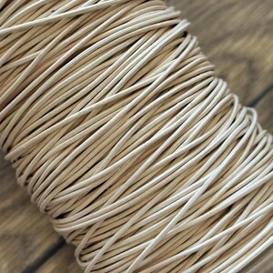 May include: Close-up of a spool of off-white, round leather cord. The cord is tightly wound, creating a dense, textured surface. The neutral colour and texture suggest its use in crafting or jewellery making.