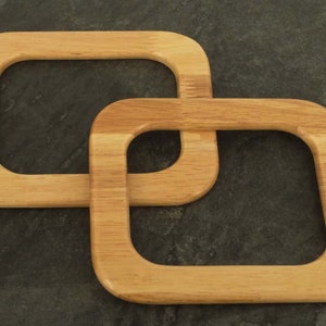 Puede incluir: Dos asas de bolso de madera clara, de forma cuadrada y esquinas redondeadas, est&aacute;n entrelazadas. Las asas son de madera clara con veta visible. El fondo es una superficie oscura y texturizada.