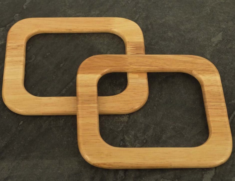 Puede incluir: Dos asas de bolso de madera clara, de forma cuadrada y esquinas redondeadas, est&aacute;n entrelazadas. Las asas son de madera clara con veta visible. El fondo es una superficie oscura y texturizada.