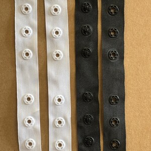 May include: Two white and two black strips of fabric with rows of plastic snaps. The snaps are white on the white fabric and black on the black fabric.