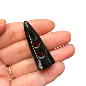 May include: A black, horn-shaped button with two holes. The button has a glossy finish and a slight curve. The button is held in a hand against a white background. The button is a sewing or craft supply.
