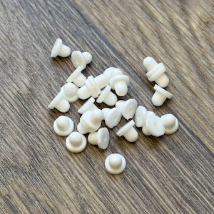 May include: A collection of small, white, mushroom-shaped earring backs scattered on a wooden surface. The earring backs have a flat, circular base and a rounded top, designed to secure earrings.