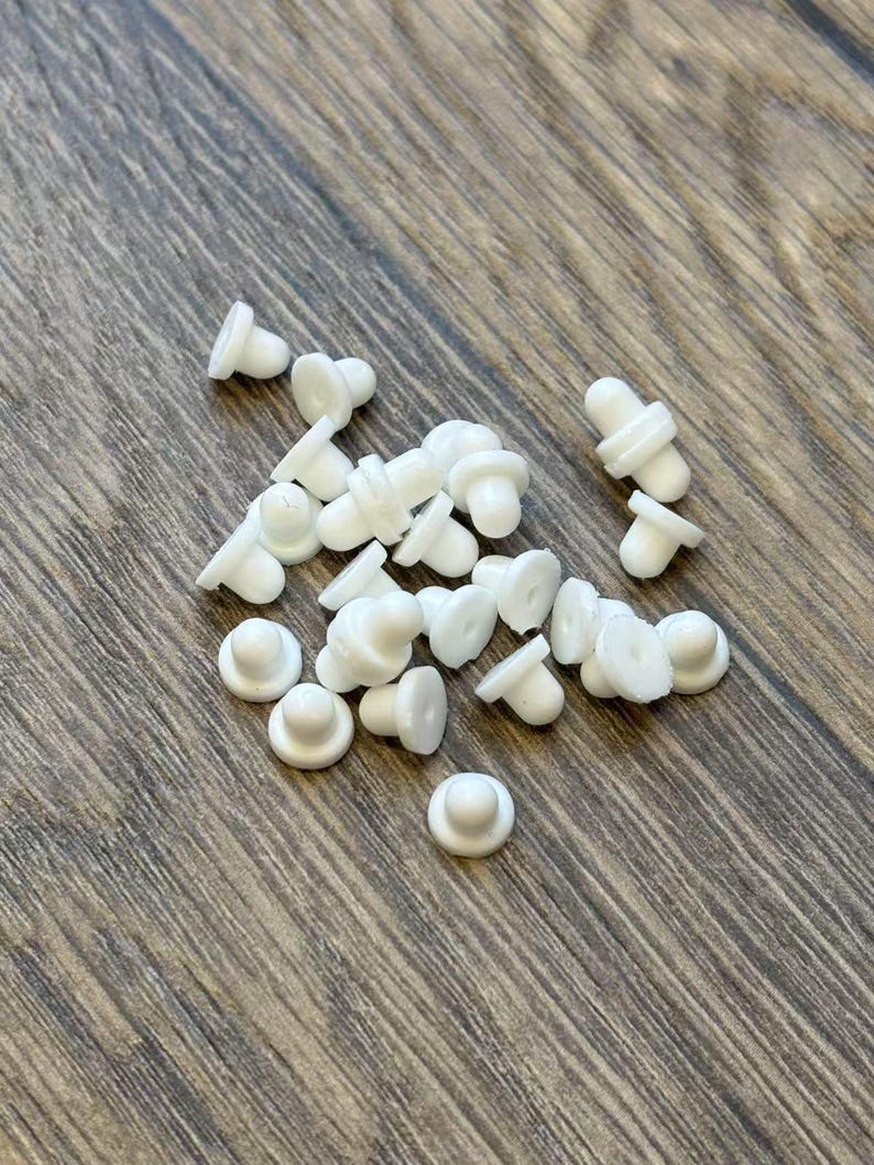 May include: A collection of small, white, mushroom-shaped earring backs scattered on a wooden surface. The earring backs have a flat, circular base and a rounded top, designed to secure earrings.