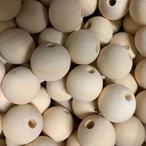 May include: A close-up of a pile of natural wooden beads. The beads are spherical, with a hole drilled through the centre. The wood is a pale, unfinished colour, suggesting they are ready for painting or other craft projects.
