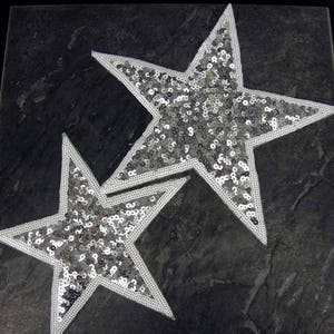 May include: Two silver sequin stars with white outlines. The stars are layered on top of each other.