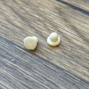 May include: Two small, white, plastic objects with a rounded top and a flat base. One object has a slightly irregular shape, while the other is more symmetrical. The objects are on a wooden surface.