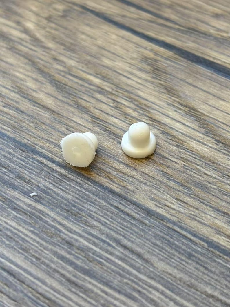 May include: Two small, white, plastic objects with a rounded top and a flat base. One object has a slightly irregular shape, while the other is more symmetrical. The objects are on a wooden surface.