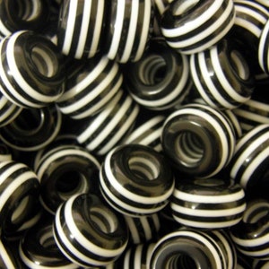 May include: Close-up of a pile of black and white striped beads. The beads are ring-shaped with a central hole, and the stripes are a consistent width. The beads have a shiny finish and are likely made of glass or a similar material.